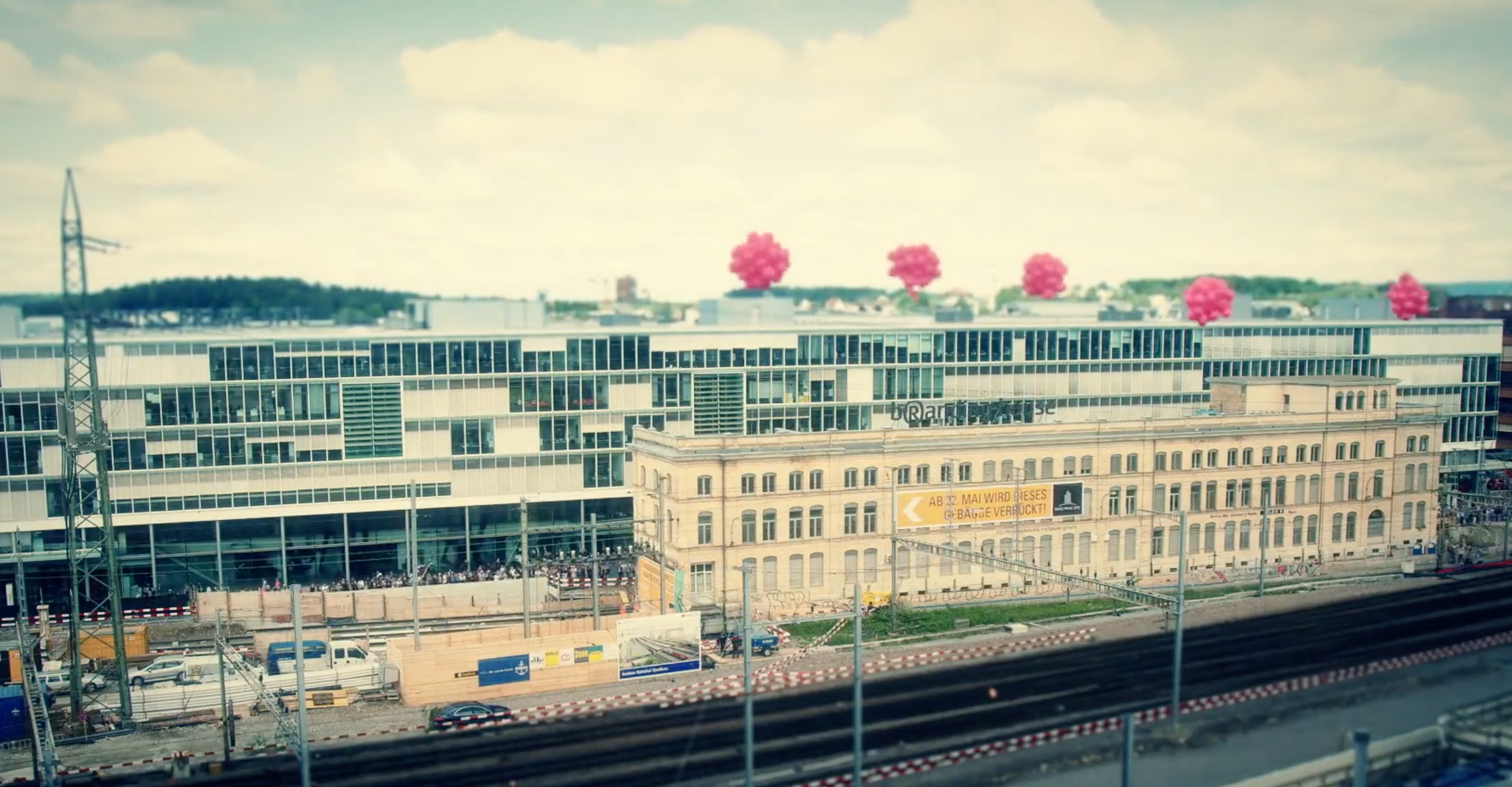 A large modern glass office building behind an older historic building with a yellow banner, situated next to train tracks with several clusters of pink balloons floating above the rooftops.