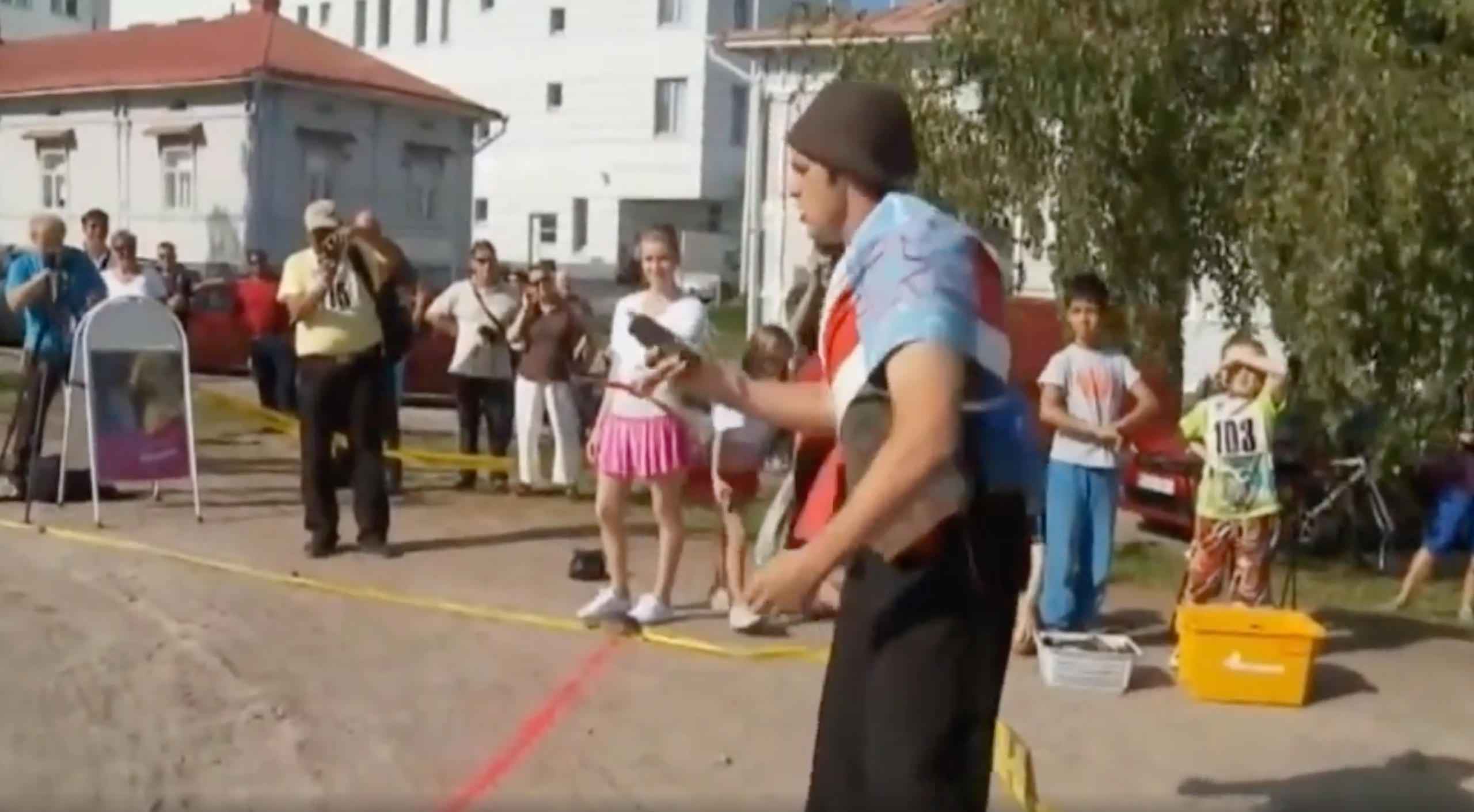 A street performer in a hat and vest juggles objects for a small crowd of spectators gathered on a paved area.