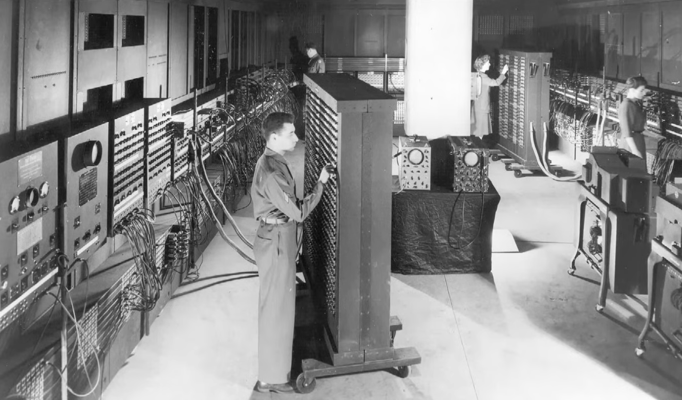 A man and women operating the large ENIAC computer with panels, cables, and dials in a laboratory.