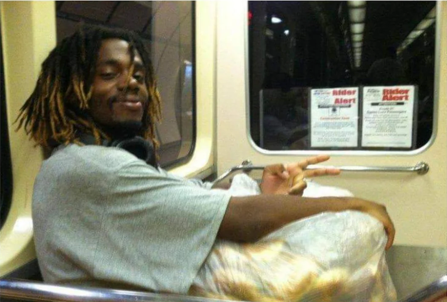 A man with dreadlocks smiles and makes a peace sign while sitting on a subway train with a large plastic bag.