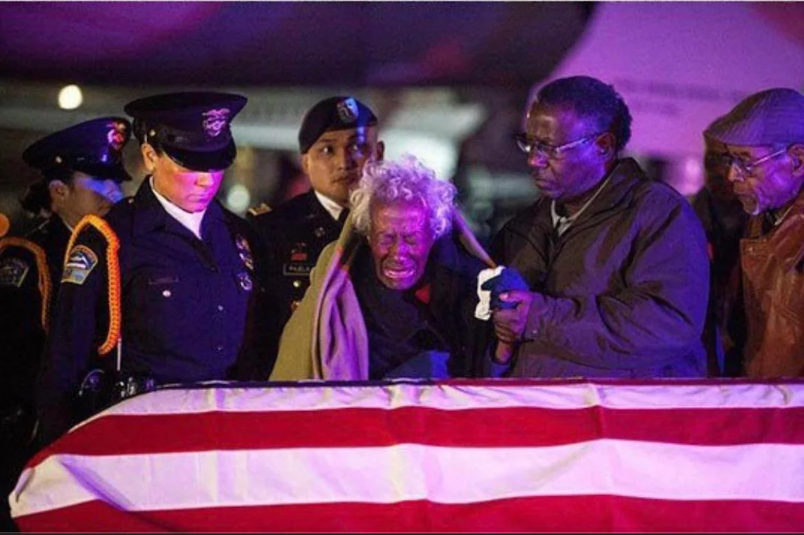 An elderly woman weeping while leaning over a flag-draped casket, supported by a man and surrounded by uniformed officers.