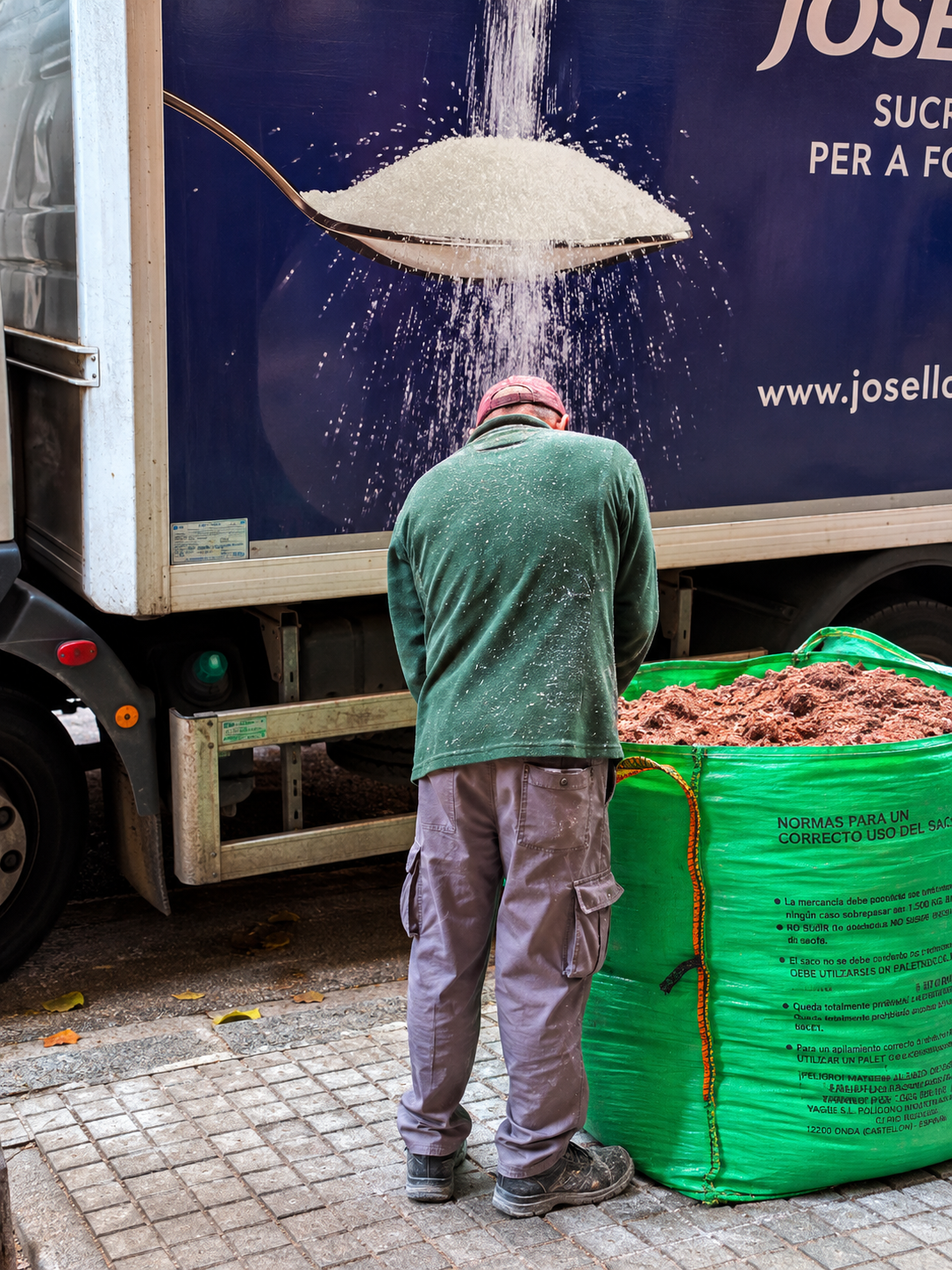 A worker in a green sweater and grey pants stands next to a large green bag filled with dark organic material, while a graphic on the side of a parked truck shows a giant spoon pouring sugar onto his back.