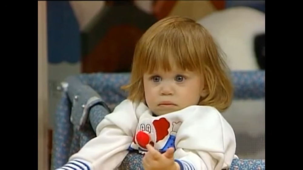 A young child with light hair wearing a clown-themed sweatshirt sits in a playpen.