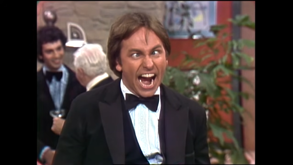 A man in a tuxedo makes a silly, cross-eyed expression with his mouth wide open.