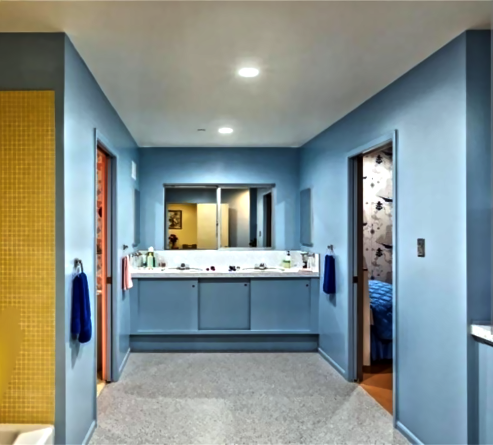 A blue-walled bathroom with a double vanity, a large mirror, and two doorways leading to other rooms.