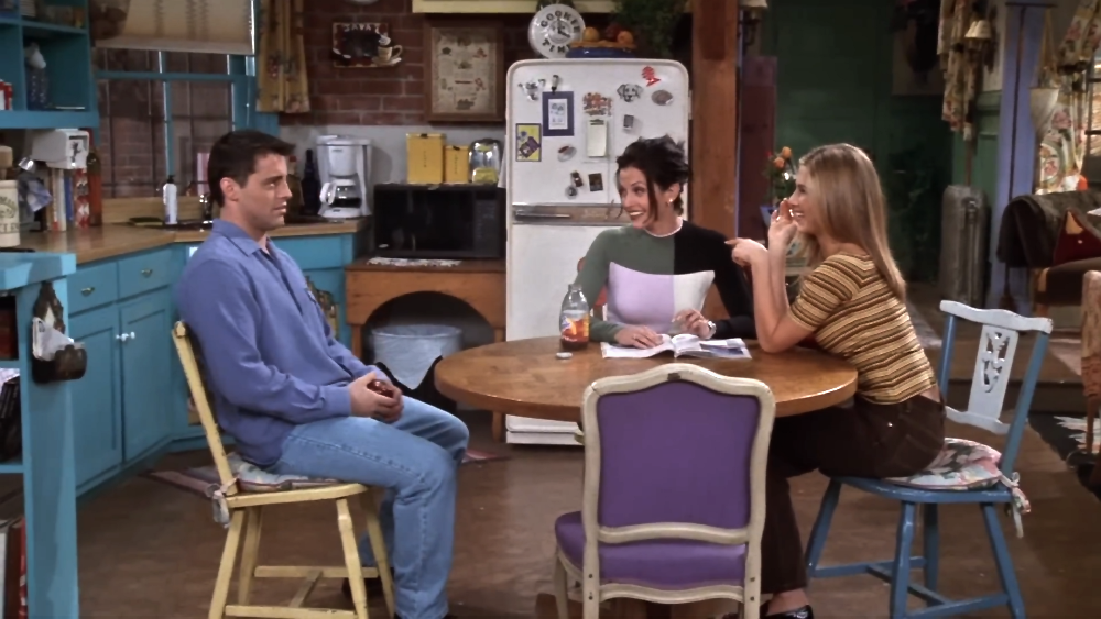 Joey, Monica, and Rachel sitting around a wooden table in an apartment kitchen.