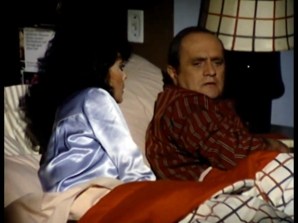 A woman in a blue satin pajama top sits up in bed next to a man wearing a striped button-down shirt.