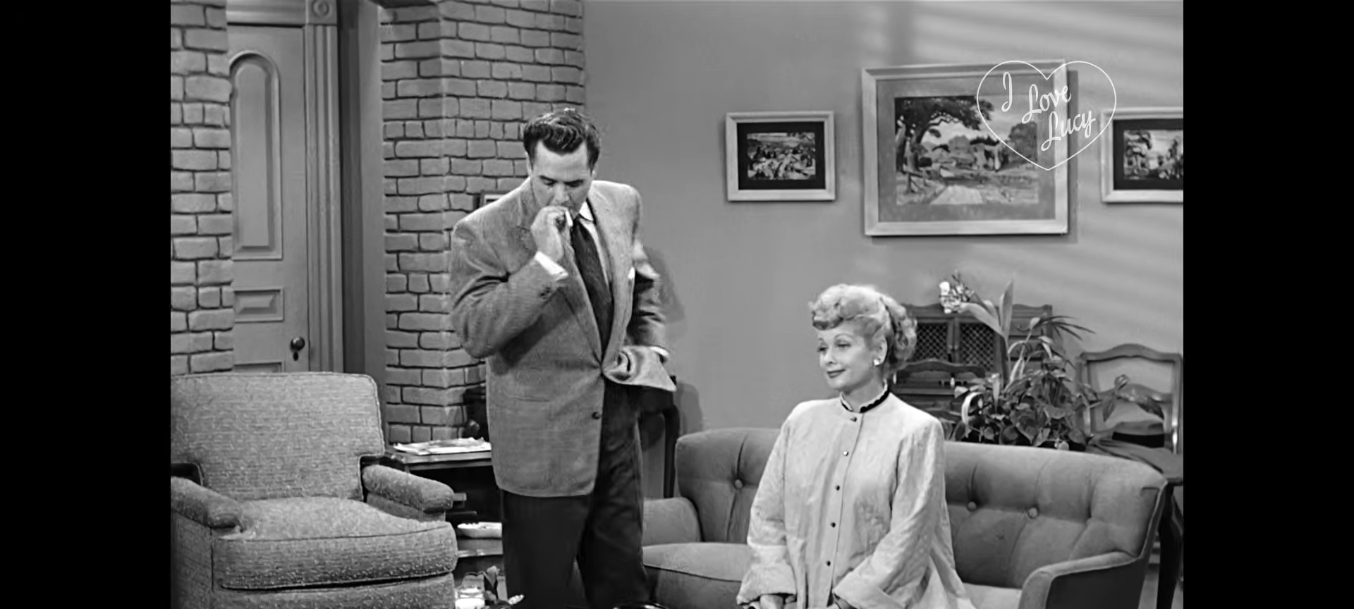 Desi Arnaz stands and looks down while Lucille Ball sits on a sofa in a scene from I Love Lucy.