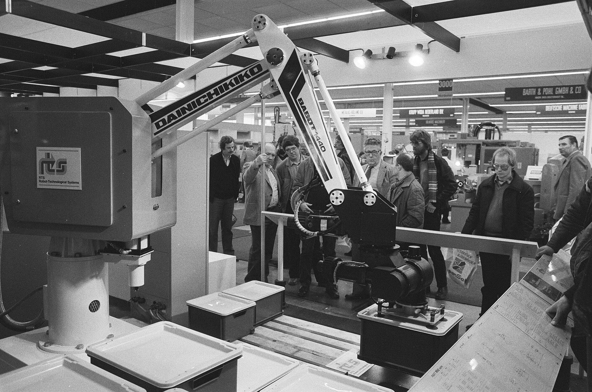 A group of people at a trade show watch a Dainichikiko industrial robotic arm operate above plastic containers on a conveyor system.