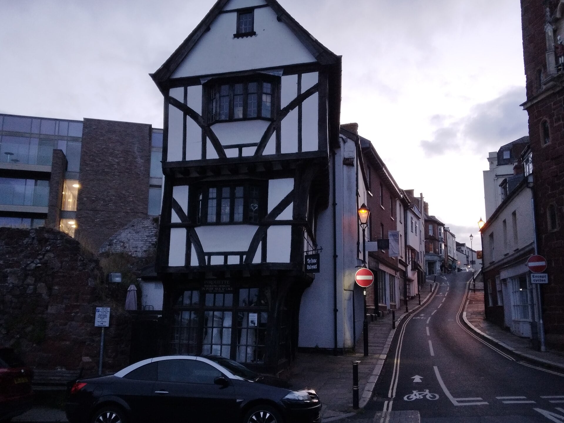 A historic half-timbered building sits on a corner at the junction of a narrow, sloping street in the evening.