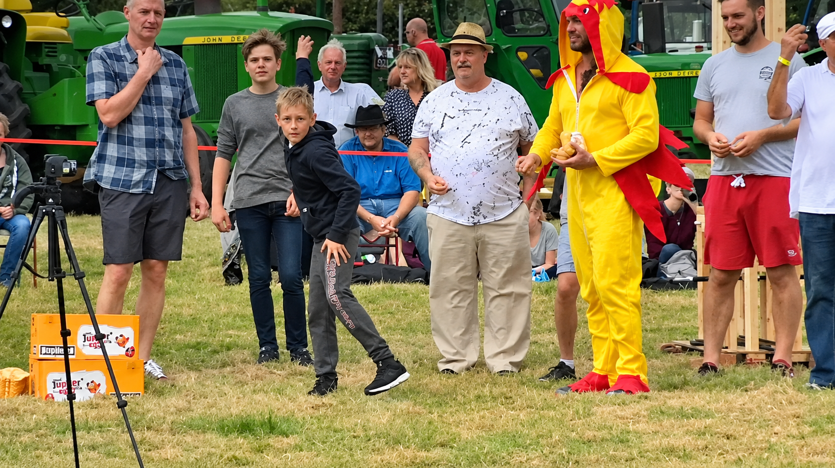A young boy in a hoodie leans forward to throw an object while a man in a red and yellow dragon costume holds a stuffed toy, with several spectators standing in front of two green tractors.