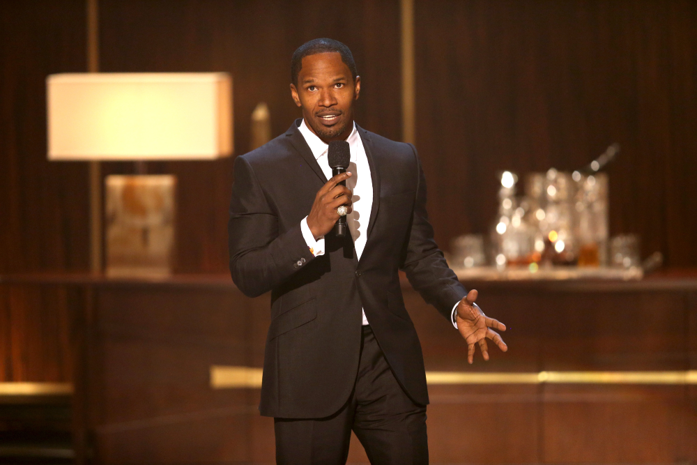A man in a black suit holds a microphone and speaks on stage, with a lamp and glassware visible on a table in the background.