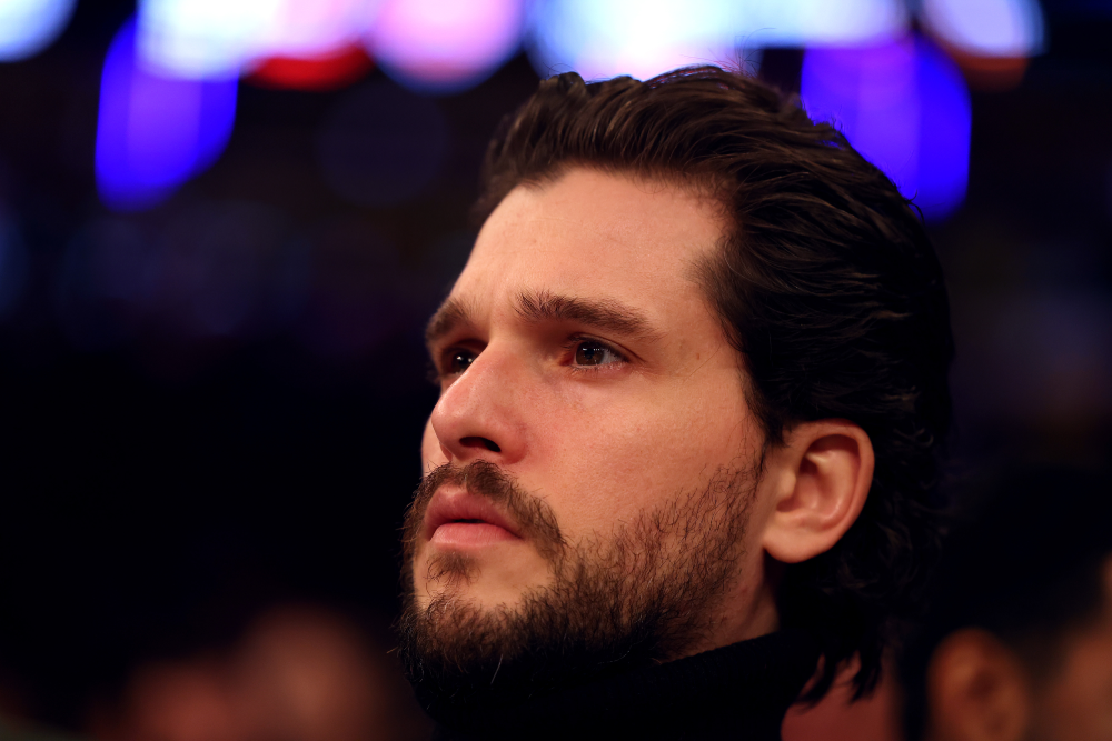 A man with a beard and mustache looks upward with a serious expression. He has dark hair and is wearing a black turtleneck. The background is blurred with colorful lights.