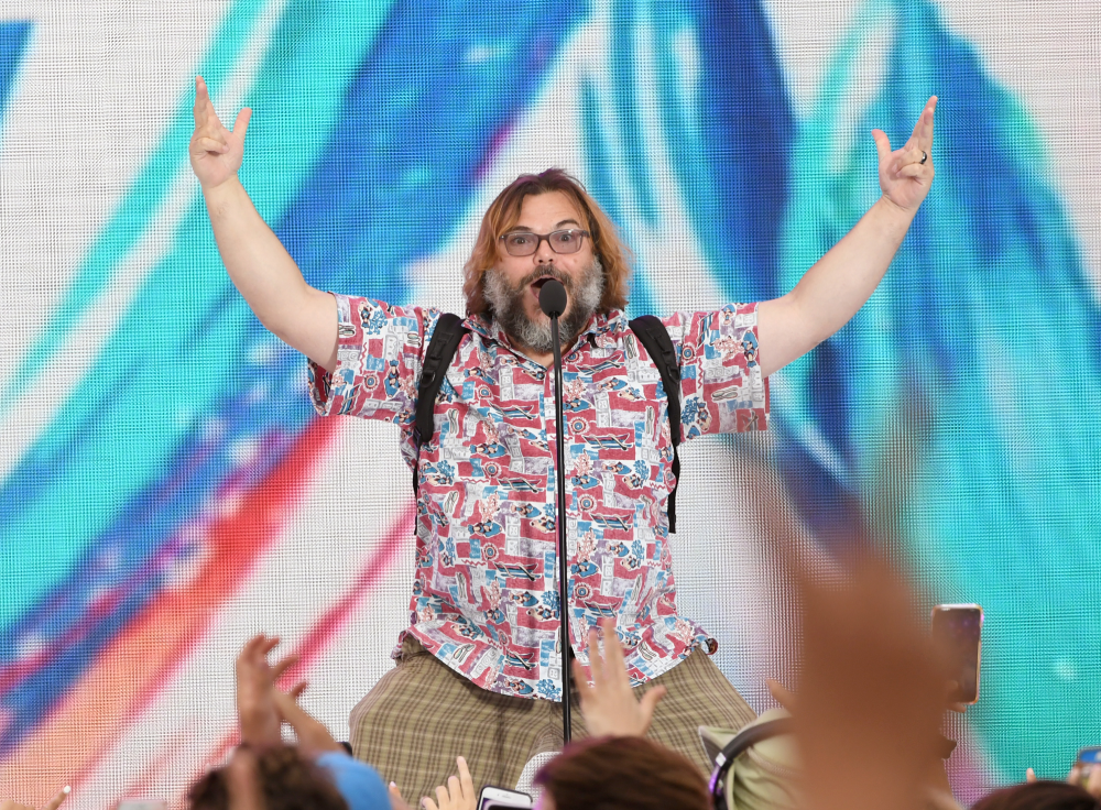 A man with long hair and a beard, wearing glasses and a colorful shirt, stands on stage with both arms raised. A crowd with raised hands is in the foreground. The background is bright with abstract blue and red patterns.