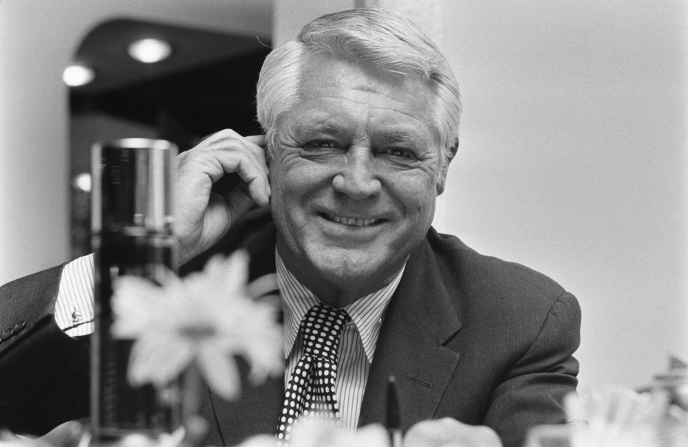 A smiling older man in a suit sits at a table, touching his ear. The black-and-white photo includes a blurred flower in the foreground.