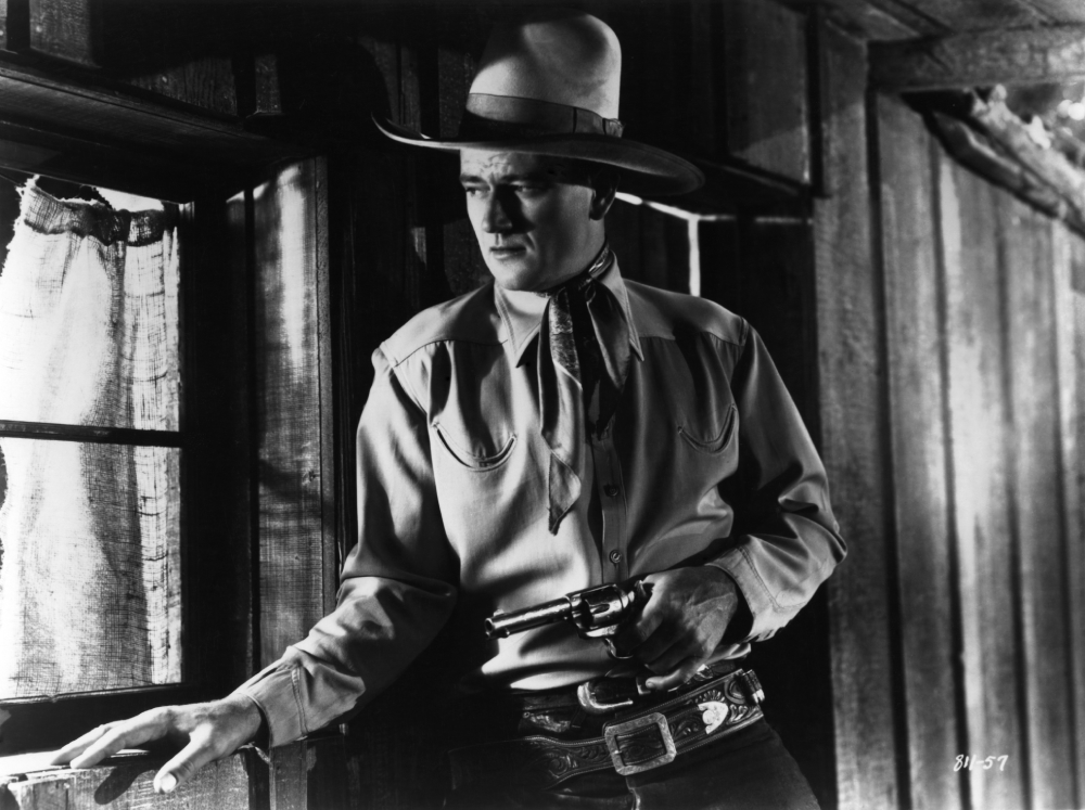 A cowboy in a hat and bandana stands by a window holding a revolver, looking outside with a serious expression. The image is in black and white and has a classic Western movie atmosphere.