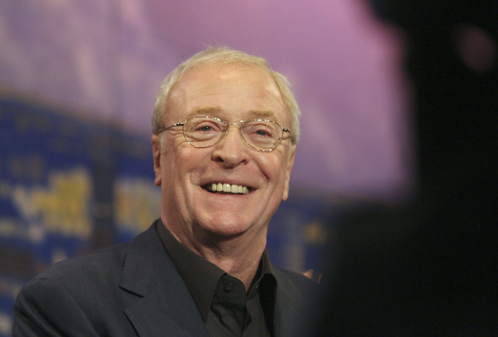 An older man with short light hair and glasses smiles warmly while wearing a dark suit jacket. The background is blurry with blue and purple hues.