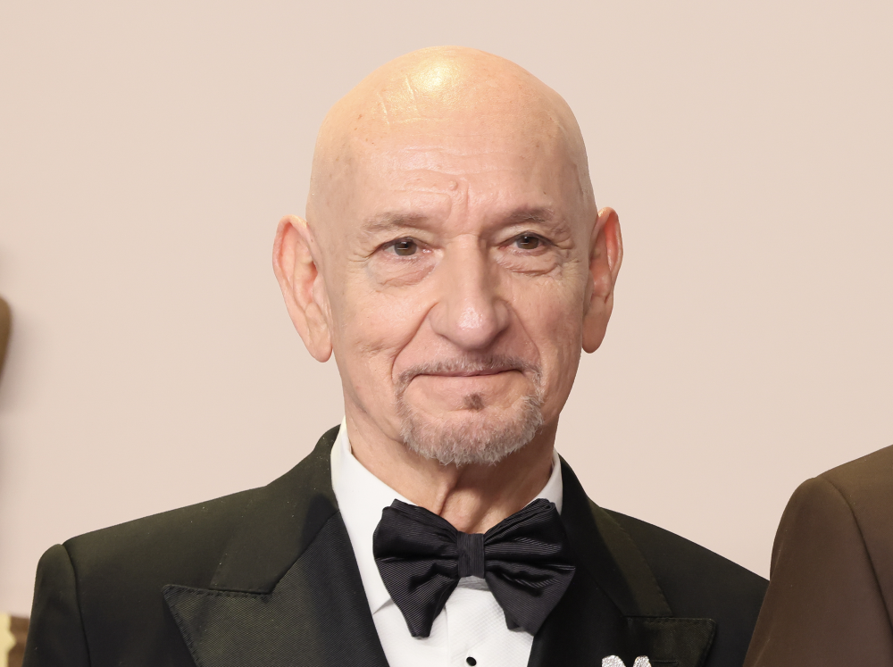 A bald man with light skin, wearing a black tuxedo and bow tie, smiles gently at the camera in front of a plain background.