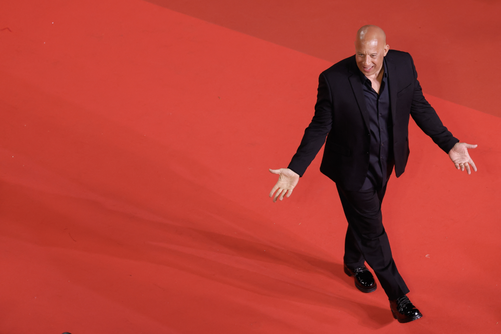 A bald man in a black suit walks confidently on a red carpet, smiling with his arms open wide.