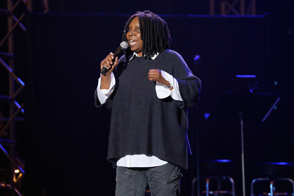 A person with dreadlocks, wearing glasses, a black poncho, and white shirt holds a microphone while speaking on stage, with a dark background and stage lights.