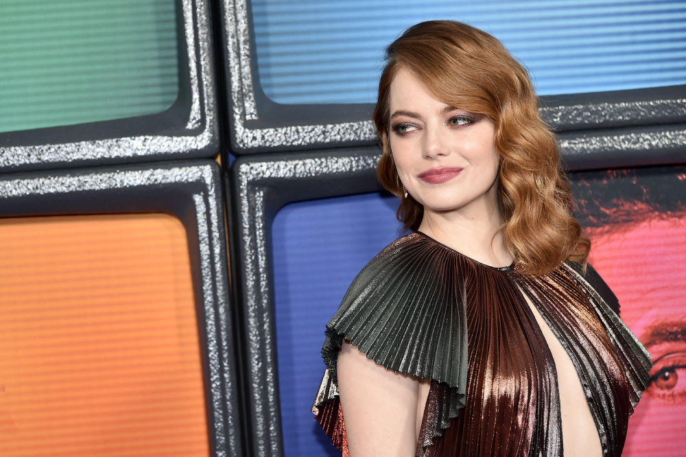A woman with wavy red hair in a metallic pleated dress poses in front of a colorful, retro TV screen backdrop, smiling slightly and looking to her right.