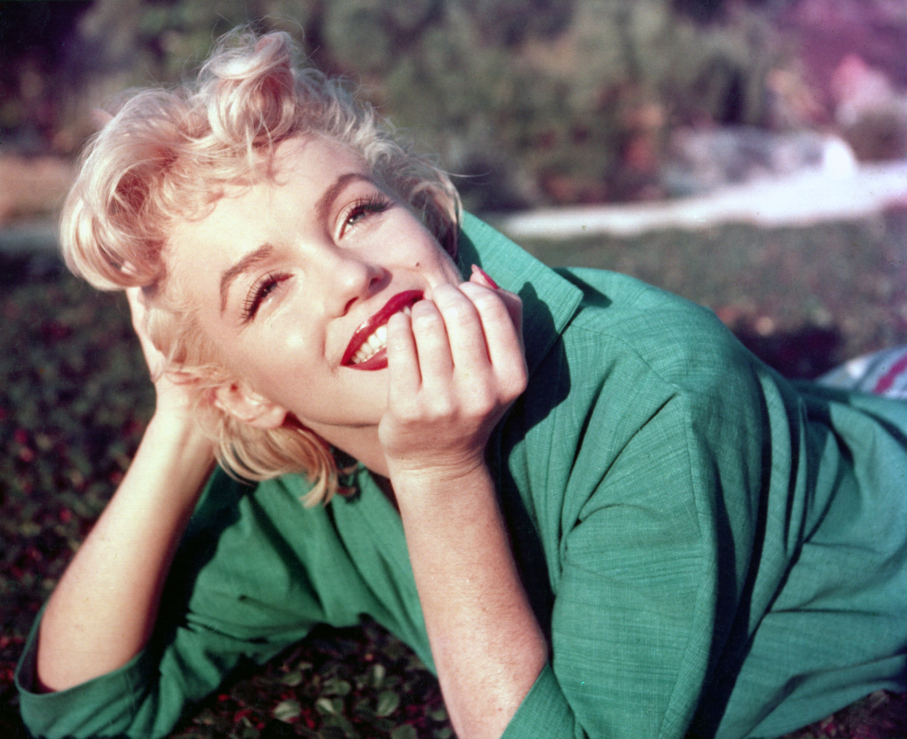 A woman with blonde, curly hair and bright red lipstick smiles while lying on her side outdoors, wearing a green shirt, with her hand resting near her mouth.