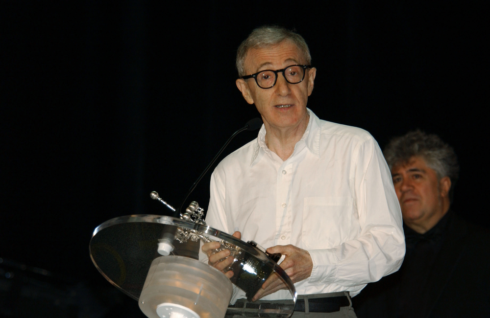 A man in a white shirt and glasses speaks at a podium, holding a small object. Another man with gray hair stands in the background against a dark backdrop.