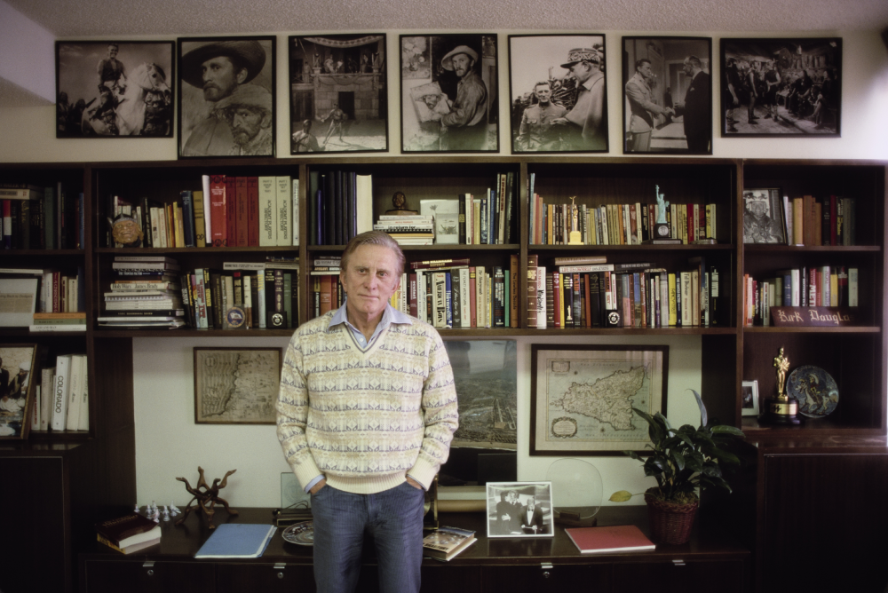 An older man stands in front of a large bookshelf filled with books, framed pictures, maps, and plants. Black-and-white historical photos are displayed on the top shelf above his head.