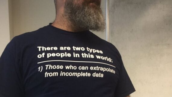 A person with a beard wears a navy shirt that says, “There are two types of people in this world: 1) Those who can extrapolate from incomplete data.”
