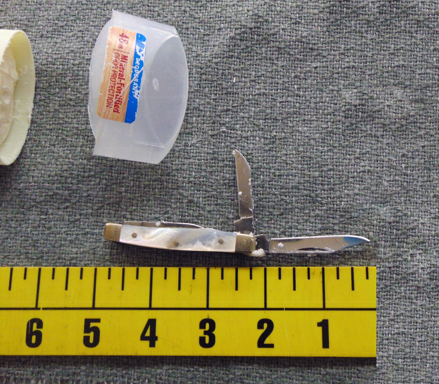 A small two-bladed pocket knife with an iridescent handle sits next to a plastic deodorant cap above a yellow measuring strip.