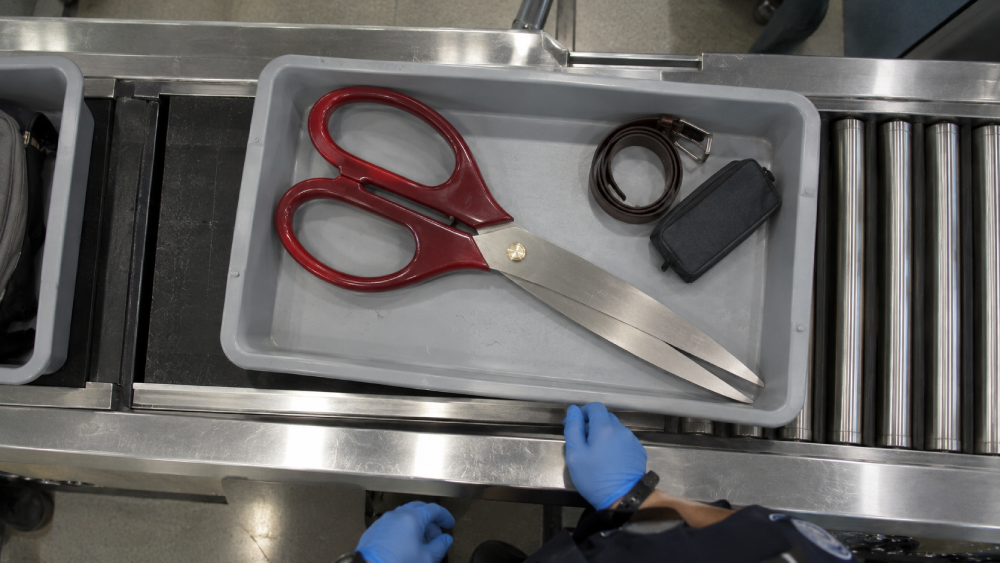A plastic tray on an airport security conveyor belt containing a pair of oversized scissors, a rolled brown leather belt, and a small black case.