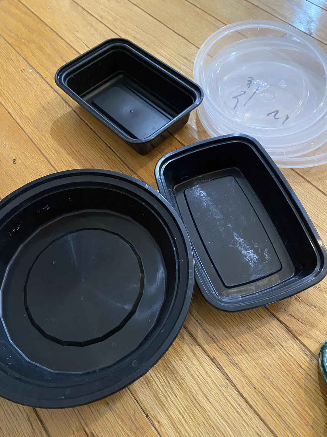 Two empty black plastic meal prep containers and one round black bowl with a stack of clear plastic lids on a wooden floor.