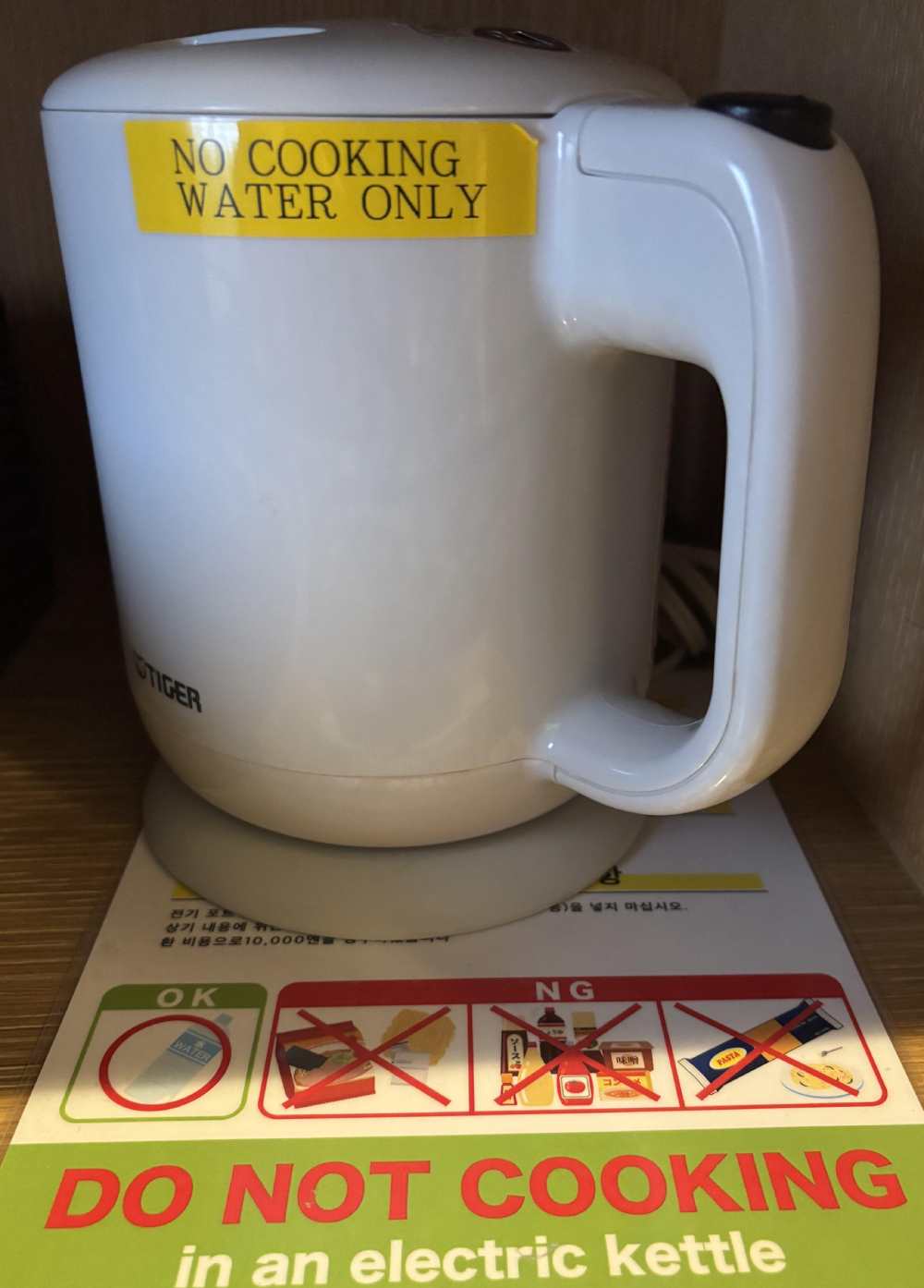 White electric kettle with a yellow warning sticker that reads, “NO COOKING WATER ONLY,” sitting on a sign that illustrates prohibited items like noodles and sauce with the text “DO NOT COOKING in an electric kettle.”