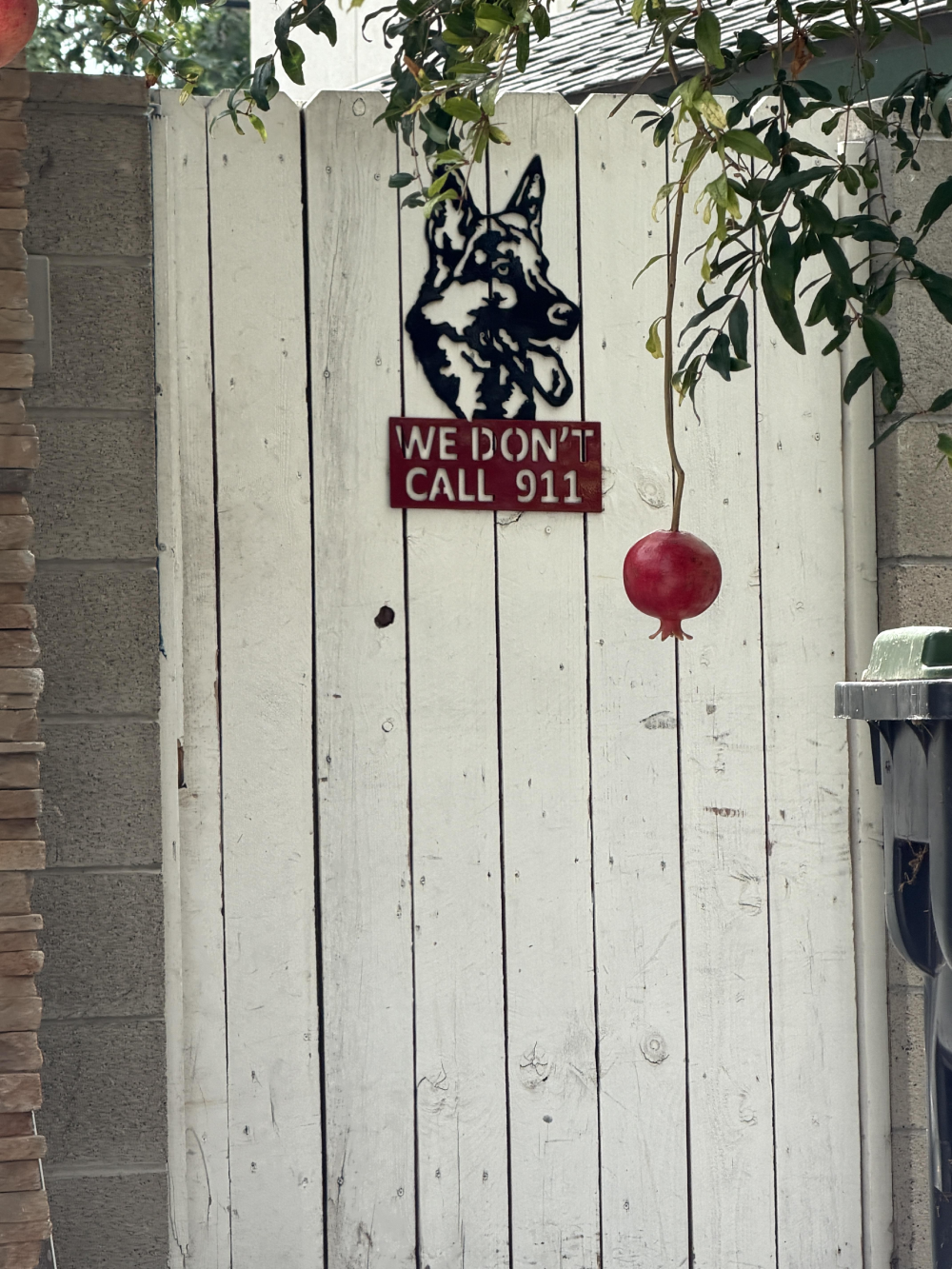 A metal sign on a white wooden gate featuring a silhouette of a German Shepherd above the text “WE DON’T CALL 911,” with a pomegranate hanging from a branch in front of it.