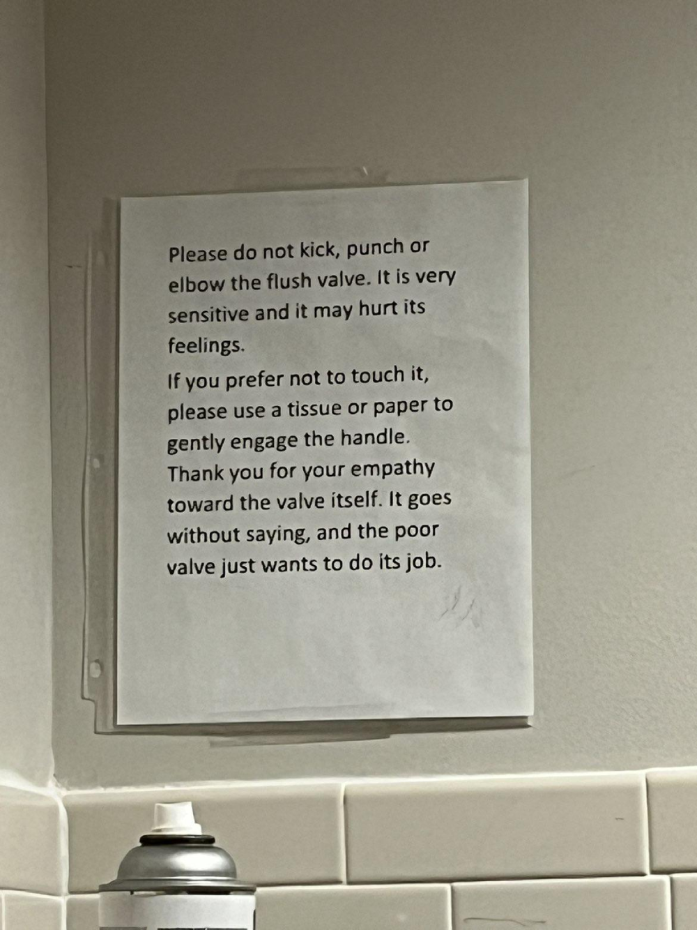 A sign taped to a wall politely asks users not to kick or punch the toilet flush valve, suggesting they use tissue instead to be kind to the sensitive valve.