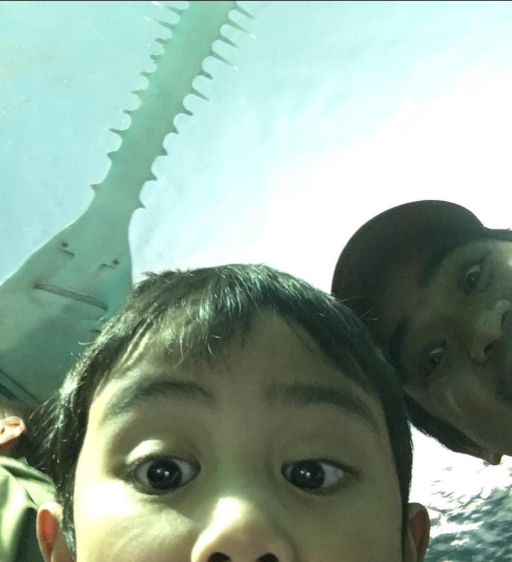 Two people take a selfie with a sawfish swimming overhead in an aquarium.