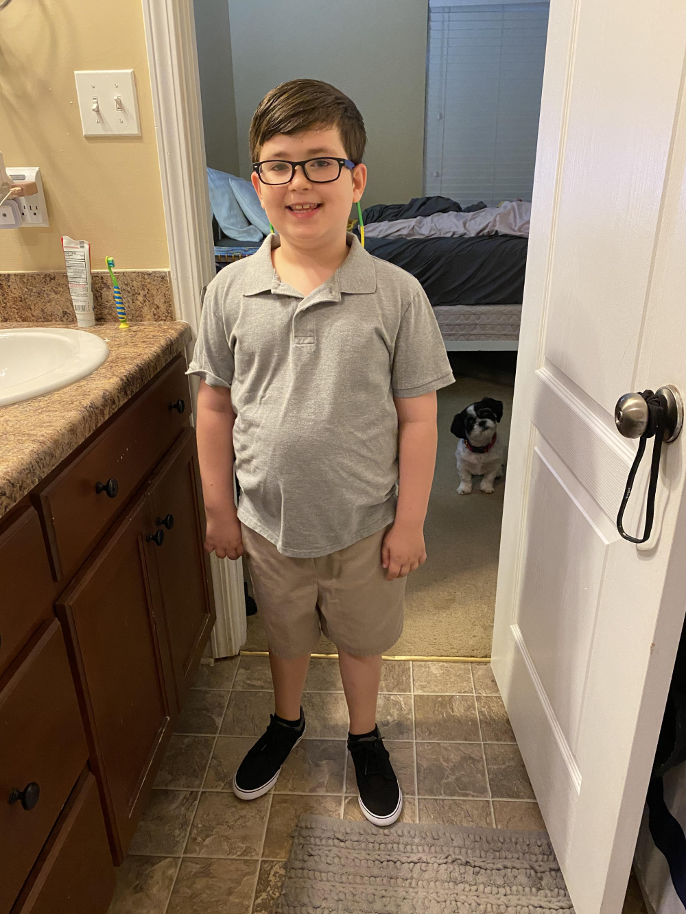 A young boy in a gray polo shirt and tan shorts stands in a bathroom, with a small dog watching from the doorway behind him.
