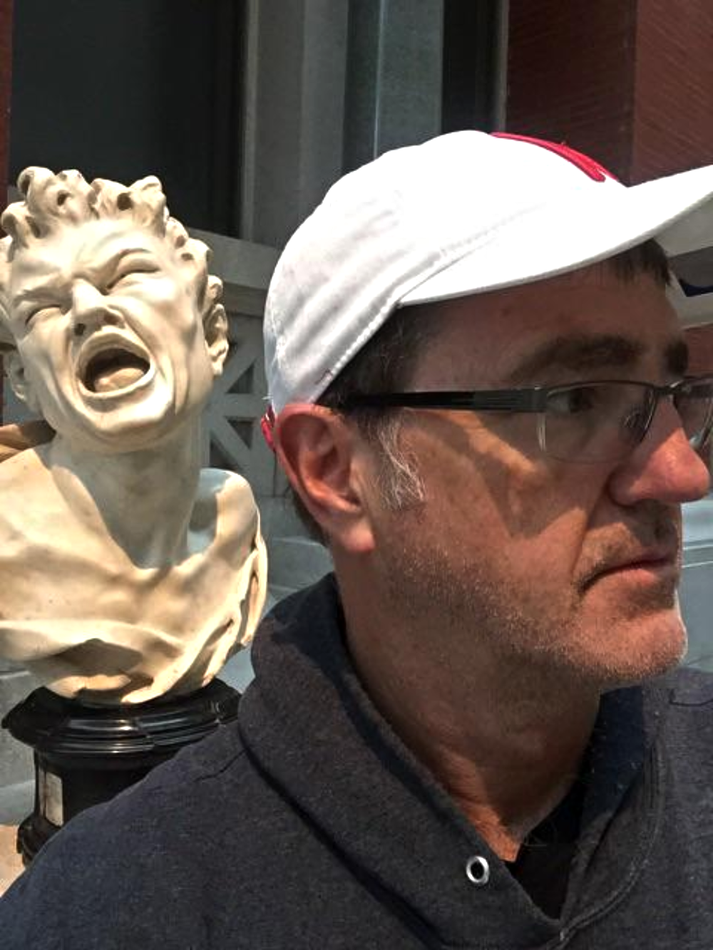 A man wearing a white baseball cap and glasses stands next to a white marble bust of a screaming figure.