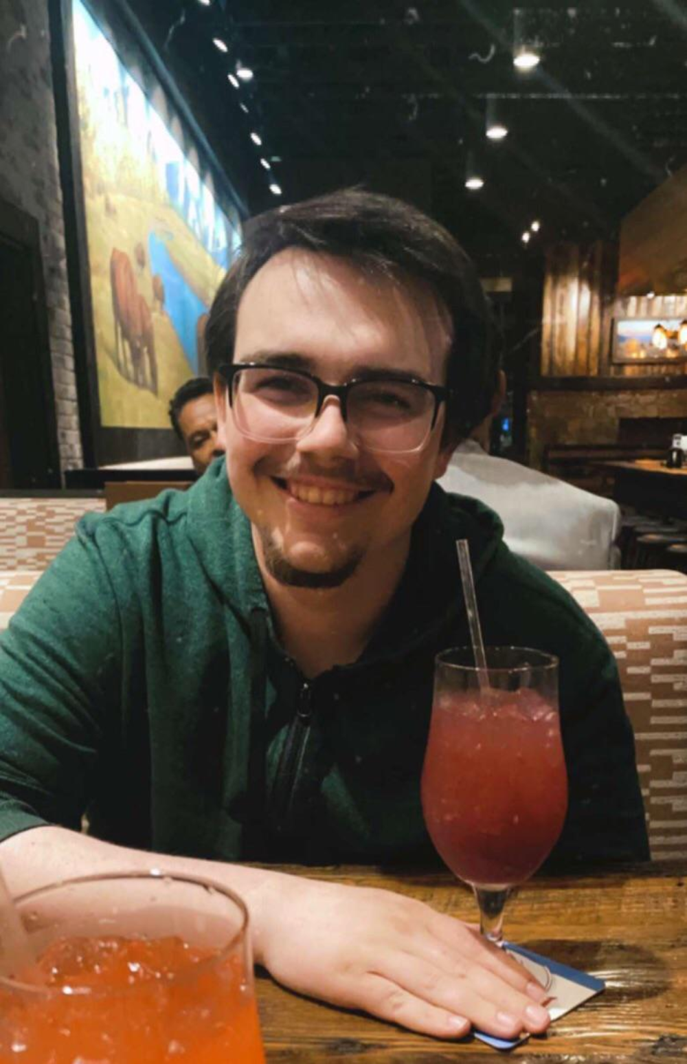 A man with dark hair and glasses smiles while sitting at a restaurant table with two red drinks.