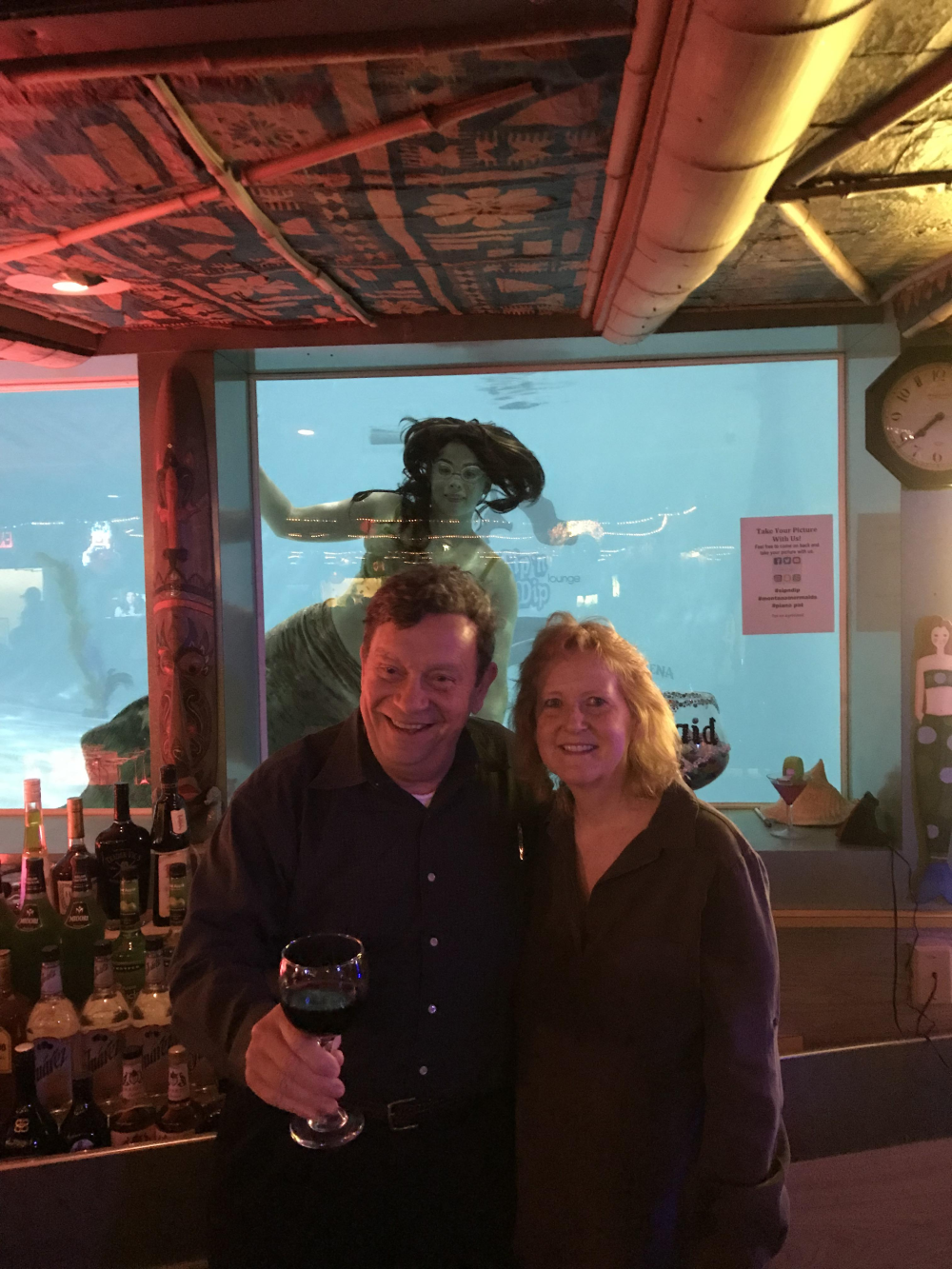 A man holding a glass of wine and a woman stand smiling in front of a large aquarium window featuring a swimming mermaid performer.
