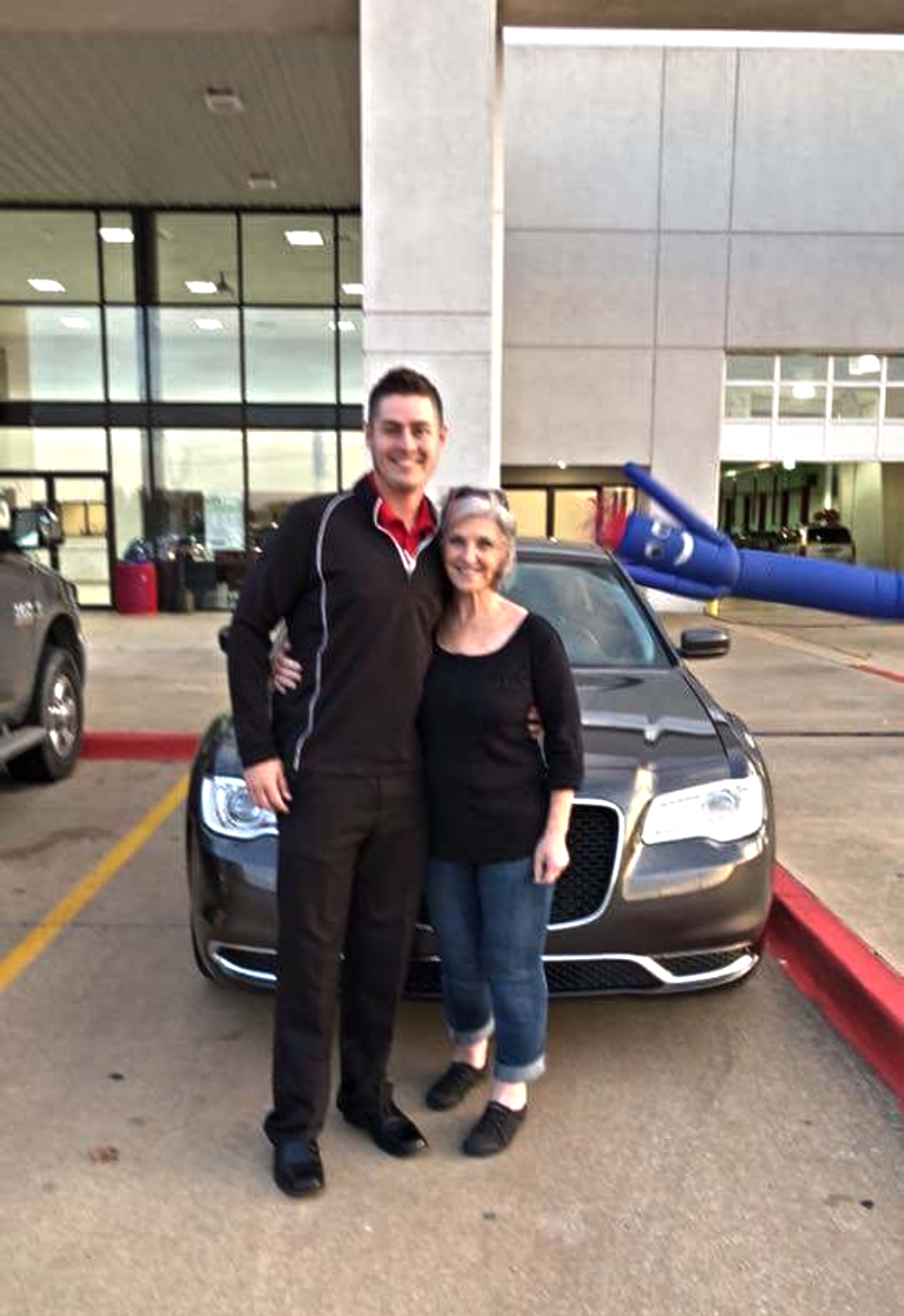 A man and a woman pose for a photo in front of a dark sedan outside a dealership, with an inflatable tube man waving in the background.