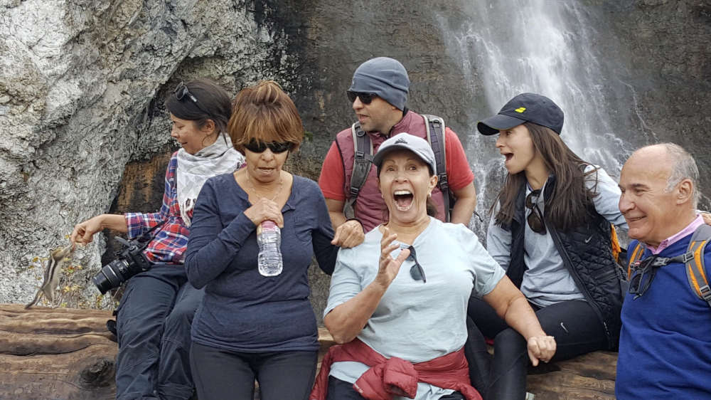 A group of six people sit on a log in front of a waterfall, with one woman cheering excitedly as a chipmunk approaches her hand.