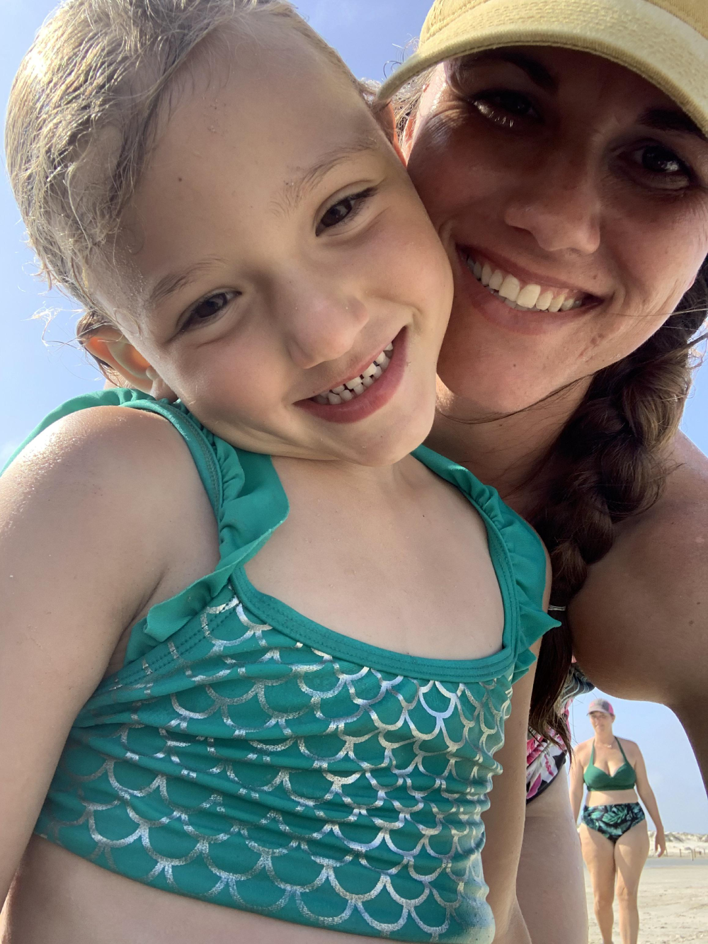 A woman in a yellow cap and a young girl in a teal mermaid-scale swimsuit smile together in a beach selfie. Another woman in a bikini walks in the background.