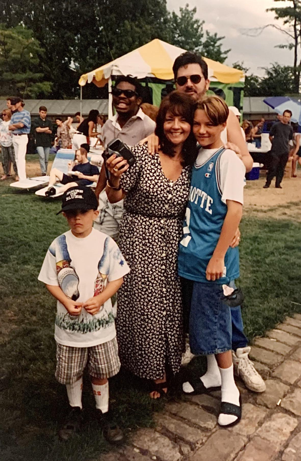 A woman with her arms around two boys outdoors, with other people and a yellow tent in the background.