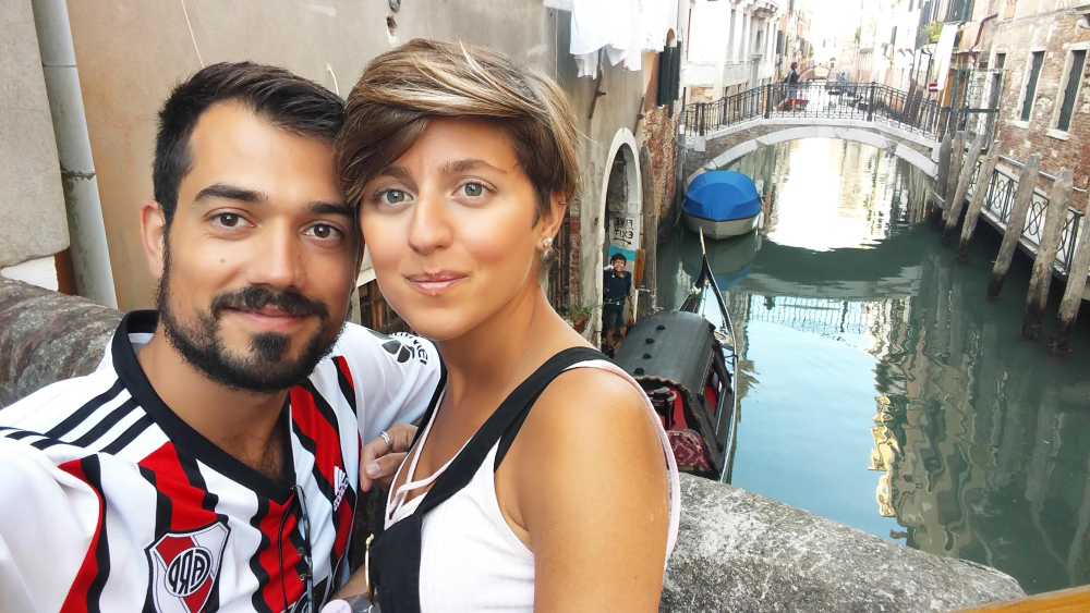 A man and woman pose for a selfie on a bridge overlooking a canal in Venice.