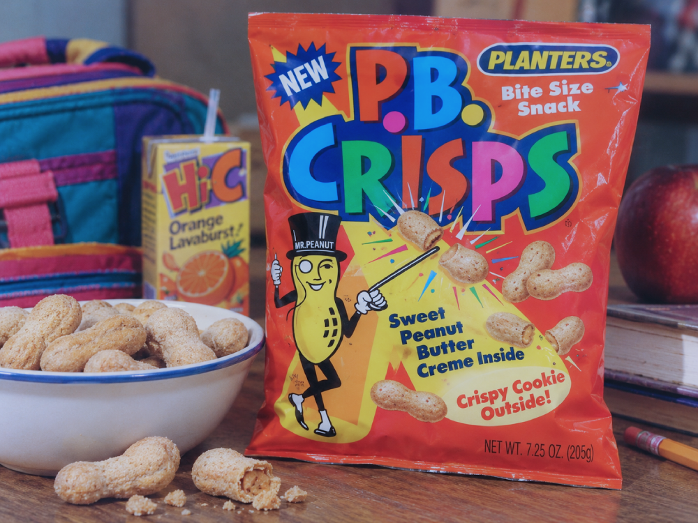 A bag of Planters P.B. Crisps sitting on a table next to a bowl of the peanut-shaped snacks, a juice box, and an apple.