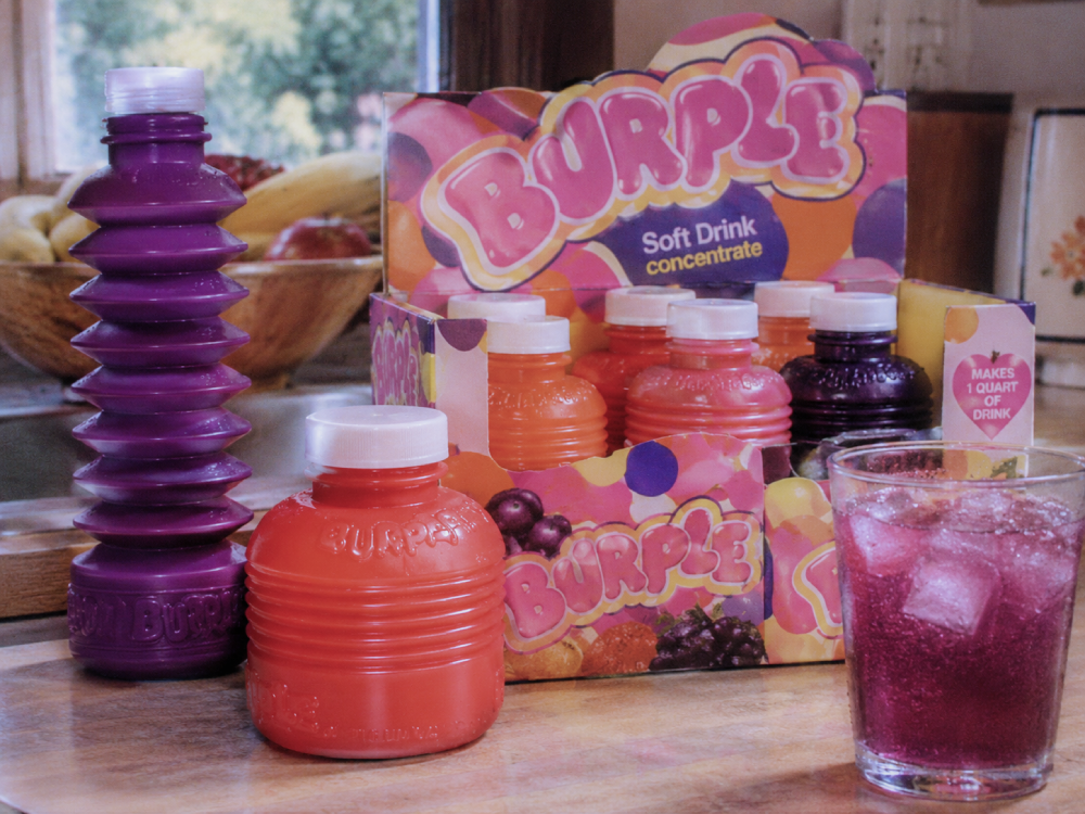 Burple soft drink concentrate bottles and a display box, with a glass of purple drink containing ice cubes in the foreground.