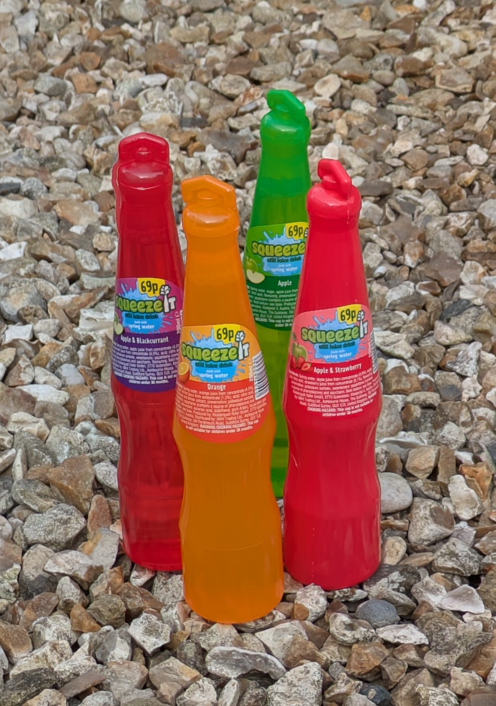 Four Squeeze-It juice drink bottles in red, orange, and green standing on a bed of gravel.