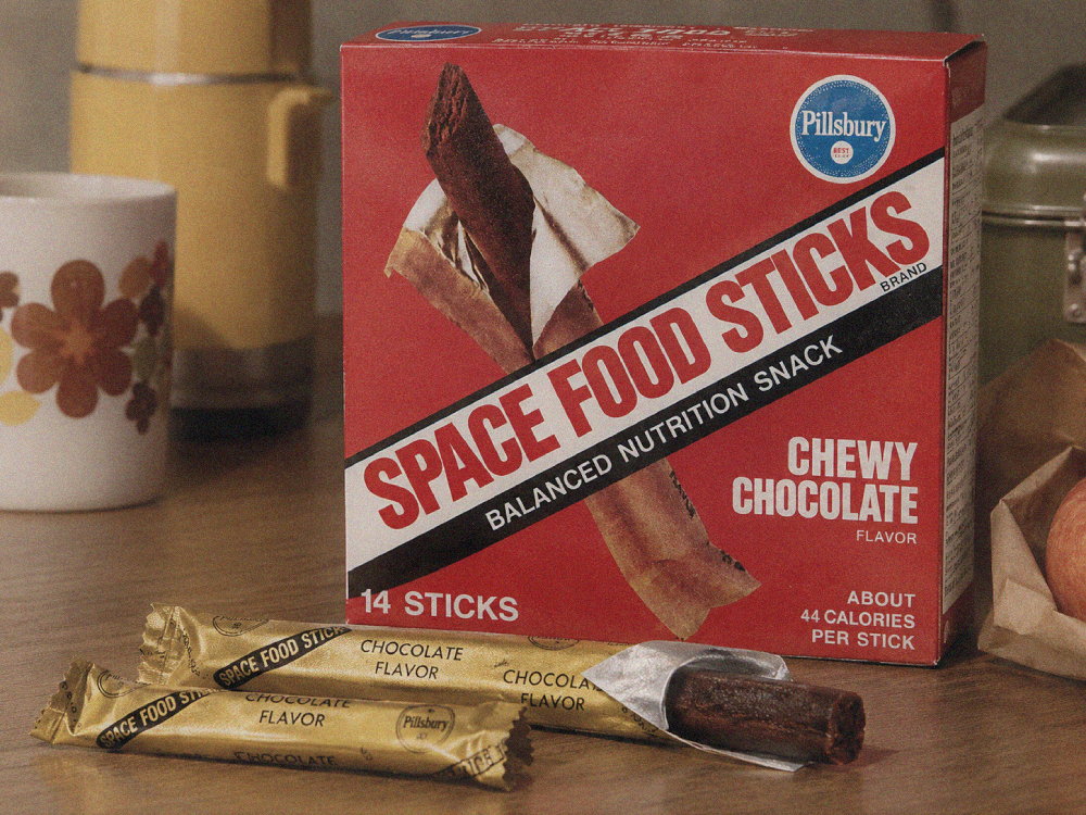 A box of Pillsbury Space Food Sticks in chewy chocolate flavor, with two individual foil-wrapped sticks on the table in front.