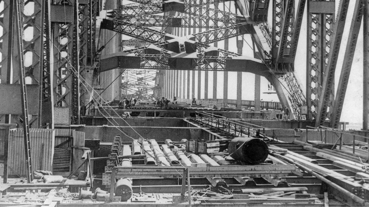 Black-and-white photo showing the construction of a large steel bridge, with beams, cables, and scaffolding visible. Workers are seen in the distance along the partially completed bridge deck.